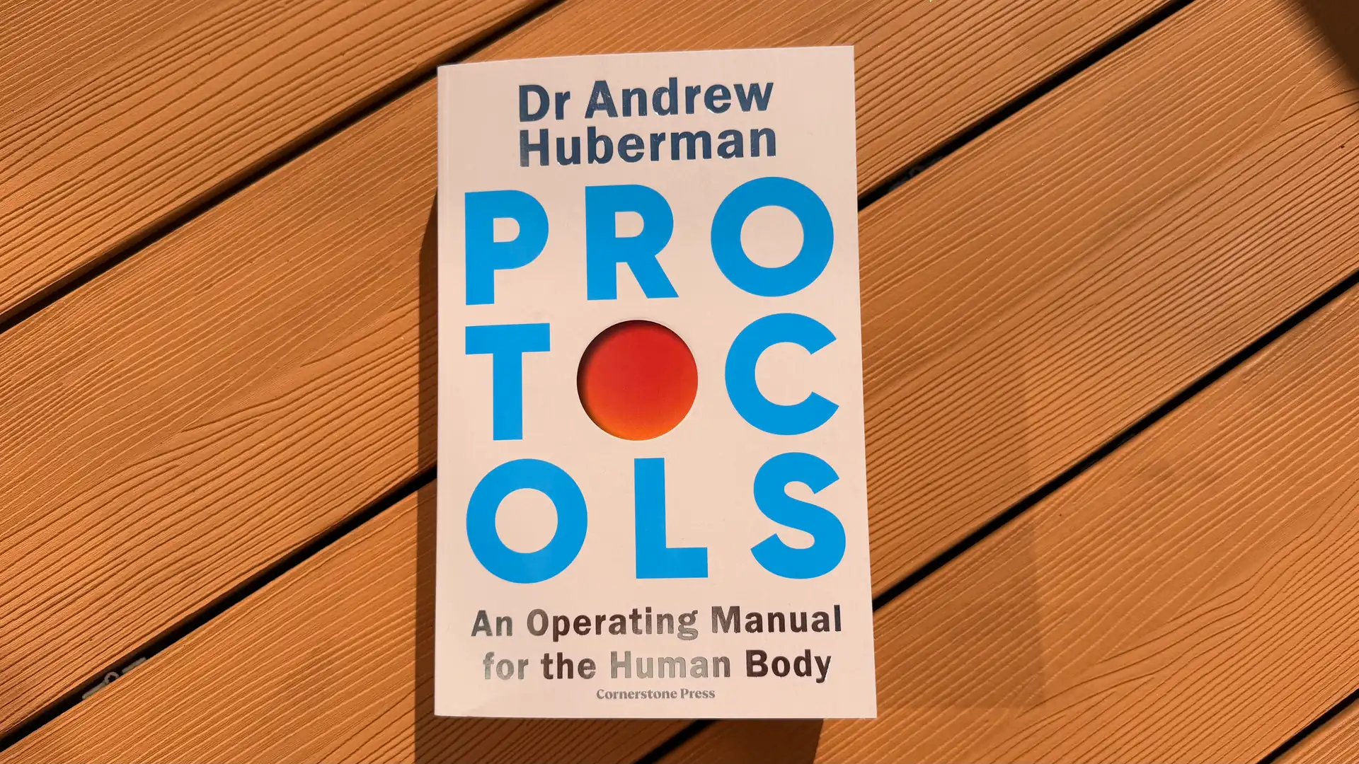 The book Protocols by Andrew Huberman with blue lettering on the cover, lying on wooden decking